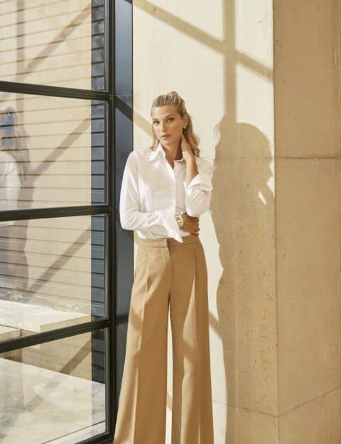Nicole Junkerman in a white shirt standing in bright daylight beside a tall window and a pale stone wall